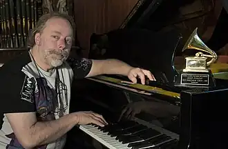 Michel Sanchez wearing a print t-shirt, sitting behind and leaning on grand piano, with right hand on keys, looking directly at camera; Grammy Award for Boheme is perched on the piano next to Sanchez's left arm