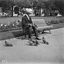 Black-and-white photograph of a young man feeding a small flock of feral pigeons while sitting on a park bench.