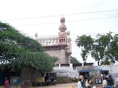 The foreground consists of a street and a small shop, a vehicle, and some people are visible. The background is a cream-colored mosque on a white platform, partially hidden by a tree.