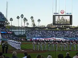 Two rows of men stand on a baseball field holding baseball caps over their hearts. A row of men in gray baseball uniforms and red caps are to the right of the image, while men in white baseball uniforms and blue caps are to the left. The stands are full with crowd members, and other people are standing in the outfield, seen in the background.