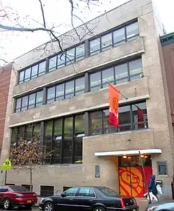 A modern-style four story building with three rows of glass windows. The front door is painted orange with a lion motif, repeated on an orange flag hanging above the entrance.