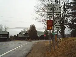 A pair of highways intersect in front of a pair of cabins in a rural area. The highway in the foreground ends here, forcing drivers to turn left or right. A nearby sign assembly indicates that NY 28 west is to the left while NY 28N east and NY 30 north are to the right.