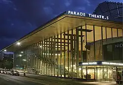 An outside, night-time shot of the building. It is well lit and has almost full-length glass panels. At the right side is the name of the drama school and also the lettering for Parade Theatres. An interior stairway is visible on the left side and a rounded wooden structure at right.