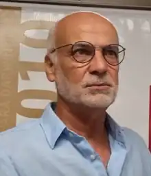 Niles Oriques, a bald middle-aged man with a white beard and glasses. He is looking away from the camera with a serious expression.
