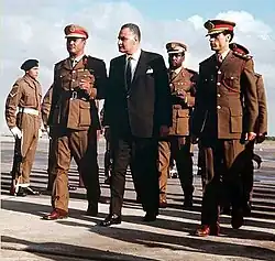Three men walking side by side. The man in the middle is wearing a suit, while the two to his side are wearing military uniforms and hats. There are a few other men in uniform walking behind them