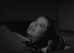 Noriko, in traditional costume, lying on a futon and covered with a blanket, stares at the ceiling, smiling