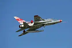 Jet fighter in a moderate left bank with hinged speed brake extended from lower fuselage.