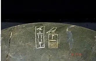 Close up of a dark stone bowl inscribed with white hieroglyphs showing an Horus falcon above a serekh and further signs on the left
