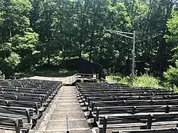 A view of the Washington Crossing Open Air Theatre in 2023. The camera is situated in the middle of the House Right seating facing the stage. The outdoor venue is showing significant signs of neglect, and part of the stage has fallen away.
