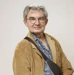 caucasian male with silver hair and spectacles wearing a royal blue floral shirt and beige jacket