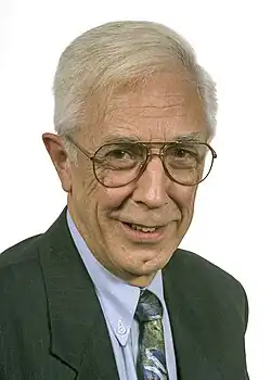 Portrait of Crampton in his sixties. He is wearing a black suit and light-blue shirt, as well as glasses and a tie including several colours. His hair is grey and combed over.