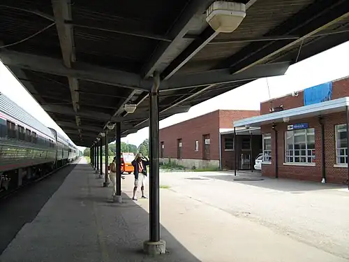 A passenger train stopped at a side platform. A brick modernist station building is at right.