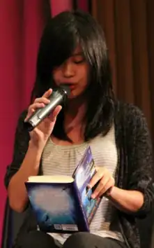 blurry image of Pieretta Dawn wearing a white top with grey stripes and a dark grey cardigan, sitting onstage, appearing to read from a book she is holding on her lap, microphone in one hand.