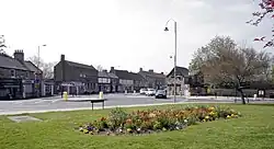 A grassed area, surrounding a large flower bed, alongside a traffic roundabout. A row of shops can be seen across the road.