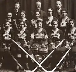 Black and white photo of hockey players wearing uniforms and equipment seated in two rows on benches