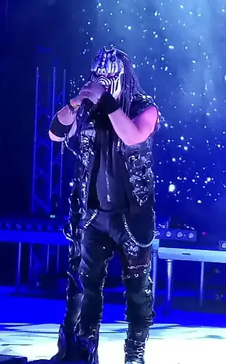 Jan Macků wearing a stylized black outfit with chains and other metal paraphernalia, with an insect-themed facemask, singing into a microphone onstge