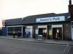 A brown-bricked building with a white sign reading "Queen's Park" in blue letters underscored by a green line and a blue line