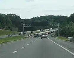 Ground-level view of a four-lane divided highway with a wide grassy median separating the opposing lanes of traffic; in the distance the road makes a sharp curve. A black electronic sign without lettering is visible above the highway.