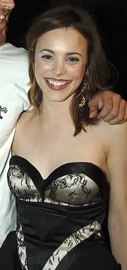 A white woman in her early 20s smiling at the camera. She has dark brown hair that reaches her shoulders, and is wearing a strapless black dress.