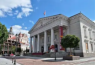 Exterior of a large, white building, with a church in the background