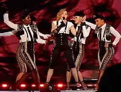 Picture of five women in front of a red background; the one in the center is blonde, has her hair in a braid and sings to a microphone. She's wearing black overalls with a white shirt underneath, and long lace gloves. The rest of the women are dressed in matador outfits.