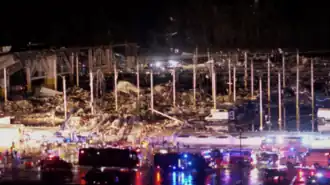First responder vehicles sit in front of the largely-destroyed warehouse in darkness, with police and fire sirens lighting up the vehicles and factory's exterior wall.