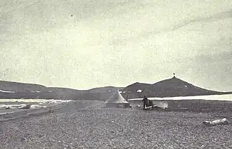 A conical tent stands on a desolate stretch of beach, with a range of low hills in the background.