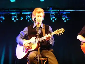 Rodney Crowell, playing an acoustic guitar while seated on a stool.