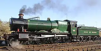 A locomotive is seen from in front and from the right. The locomotive is mainly green but the smoke box (at the front) is black and the buffer beam is red with the number 4936 written in yellow letters. The chimney is trimmed with a copper ring and the valve cover is made of brass. Behind the locomotive is a green tender with the words "GREAT WESTERN" written in yellow. In the background to the left is a signal and an engine shed with a tank engine, and to the right is a car park behind a wooden rail.