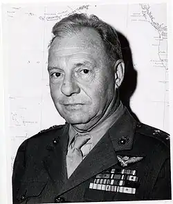 A black-and-white photo of Ross E. Rowell, a white male with short hair, wearing his Marine Corps service "A" uniform. There is a map in the background behind him. He is wearing a light-colored collared shirt and tie, with a dark jacket. His military ribbons are on the left breast side of his uniform directly above the pocket and he is wearing 2-star general rank insignia on each shoulder of his uniform jacket and dark eagle globe and anchor emblems on this jacket lapels. Directly above his ribbons, he is wearing his Naval Aviator wings.