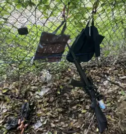 Photograph of a black rifle, digital camera, reusable shopping bag, dark backpack against a chain-linked fence