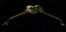 Forward shot of a starship in the black of space with distant stars in the background. The ship is primarily one color, save for a decorative pattern on its underside. The center portion of the ship is bulbous, with thin, down-stretched wings capped by long guns.