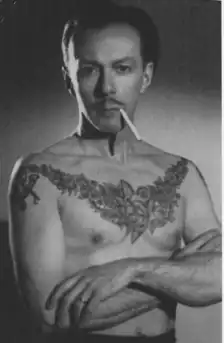 Black-and-white photograph of a shirtless, tattooed, mustachioed man with a cigarette in his mouth