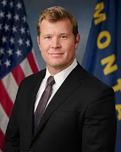 Official Senate portrait of Sheehy smiling in front of the U.S. and Montana flags, wearing a black suit, white shirt, and floral red tie.