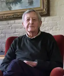 Headshot of Simon Groot. He is an elderly white man, wearing smart casual dress, sitting in an armchair in a domestic setting.
