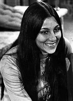 Cher smiling with long, straight dark hair parted in the center, wearing a light-colored top with buttons and a beaded necklace.