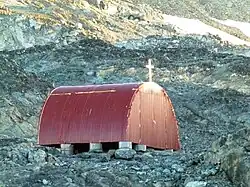 A small red Quonset Hut sittign on a concrete and metal pilings and topped with a cross sits among grey rocks.