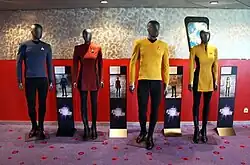 Four mannequins featuring costumes: a blue tunic with black pants, a red dress, a gold tunic with black pants, and a gold dress