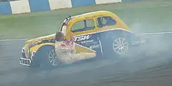 A yellow and blue car spinning on a dirt track with a white man in overalls hanging from the side door looking to the rear.