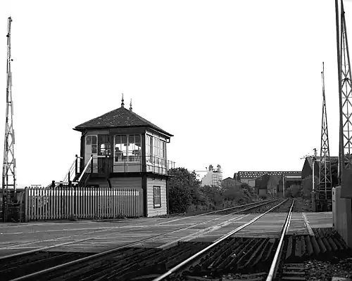 Raised cabin on left of railway lines stretching into distance on a left curve with large commercial buildings behind