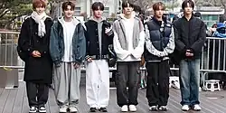 Six men standing in a row outdoors dressed in stylish winter clothing. They are wearing oversized jackets, puffer vests, hoodies, and scarves, paired with baggy jeans or wide-legged pants.