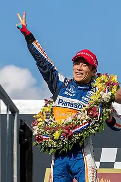 Takuma Sato in victory lane holds up two fingers indicating his two wins.