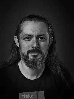 Bust length photo of man with beard and long, dark hair. He is wearing a T-shirt.