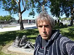 The full image of the older man in a knitted sweater displayed in the infobox: more of the background is shown, which includes the parking lot, trees, and a prominent concrete statue of two children, with each reading a book