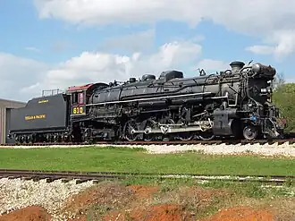 A black steam locomotive with a 2-10-4 wheel arrangement (two leading wheels, ten driving wheels, and four trailing wheels) and its tender sitting outdoors, with a shop building in the background