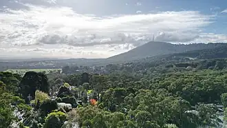 A photo of a mountain range with three radio towers atop them, trees coat the below valley in green.