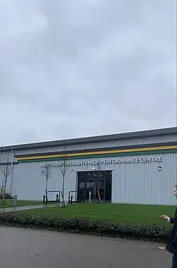 The newly opened High Performance Centre can be seen with grass in the foreground. The centre resembles a warehouse with the Saints' club colours forming a stripe along the side. In the background, the sky is grey.