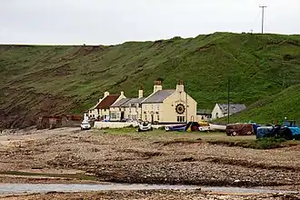 A collection of small buildings in front of a cliff, with a pebble beach in front