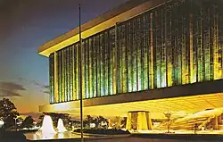 Color photo of the pavilion. The building is to the right, suspended above a plaza. There is a fountain to the left.
