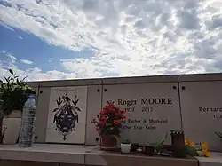 In the middle of a row of white vertical memorials, each about 1 metre wide, and with a horizontal marble shelf before each, a marker with "Sir Roger MOORE", the date "1927–2017", and "Loving Father & Husband", below which is "Our True Saint". Several flower pots and candles are on the shelf for Moore and his neighbours. To the left of Moore's marker is one with a stone plaque labelled "Moore Family", with a coat of arms in blue with gold.